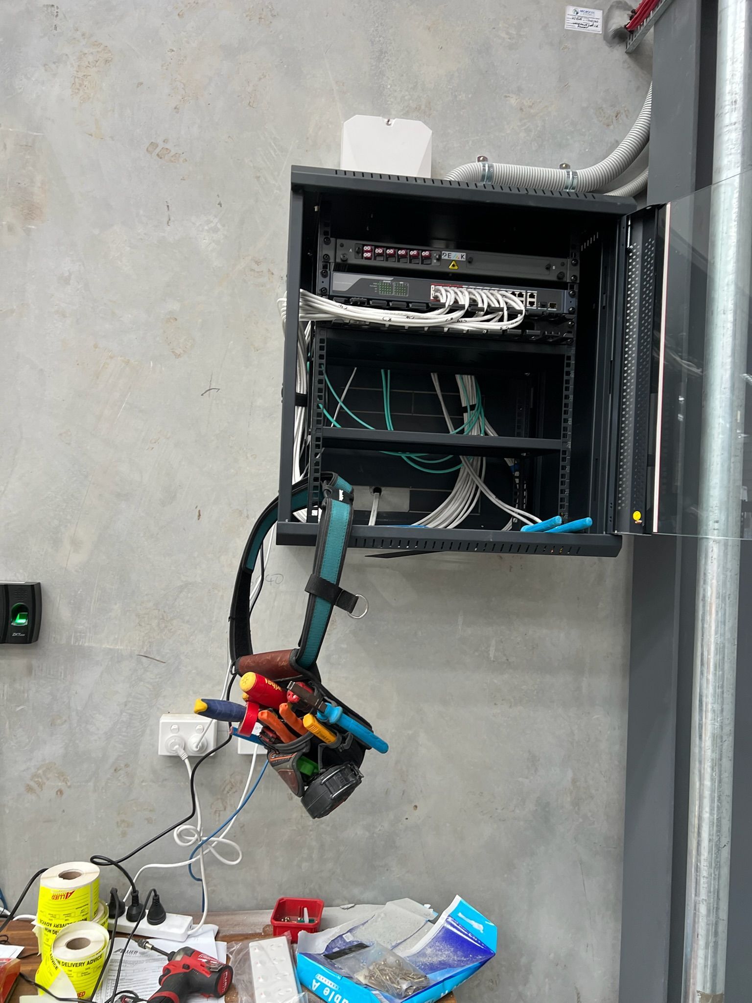 Open wall-mounted electrical panel with hanging wires and tools in a gray utility room