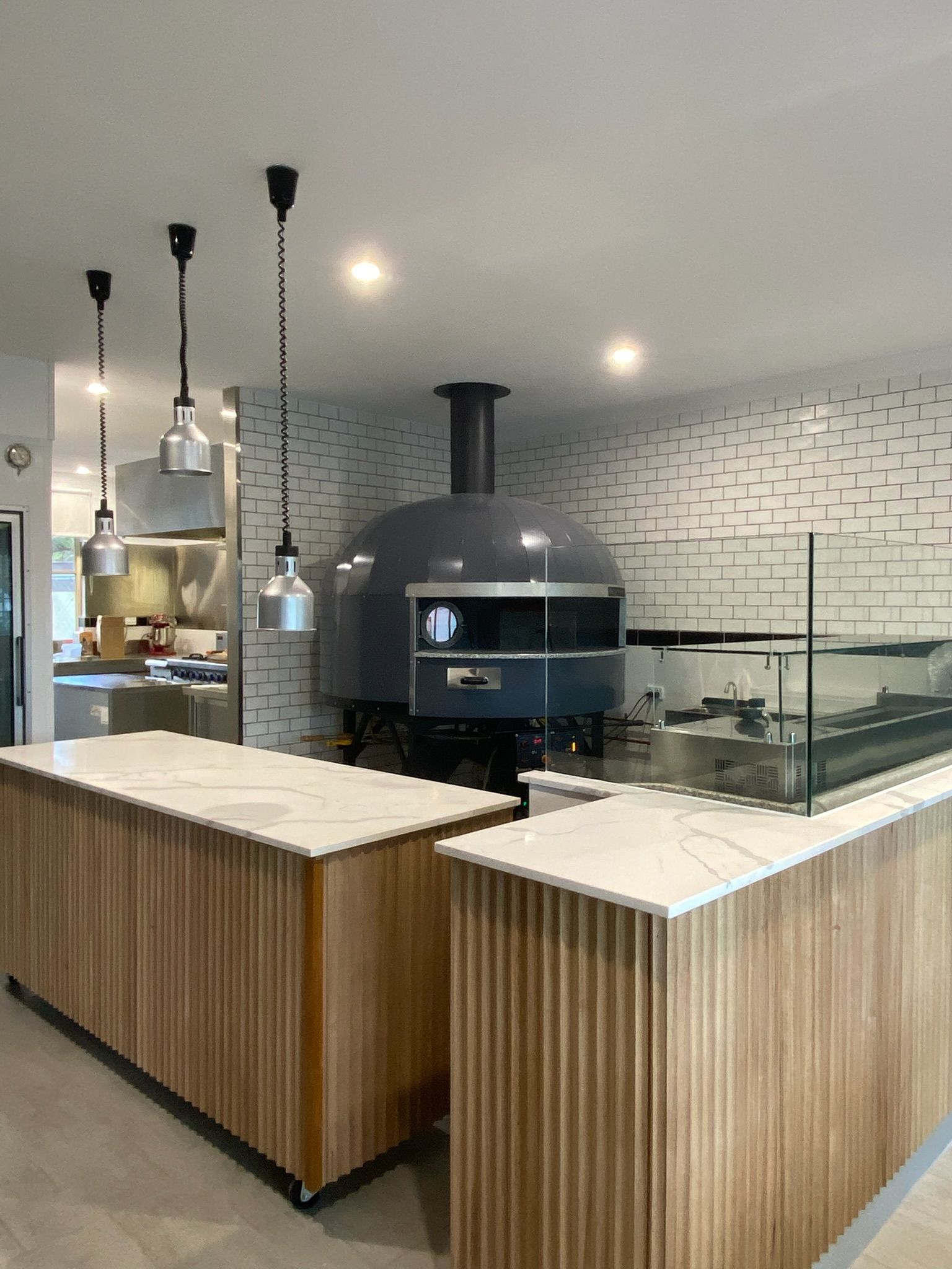 Modern café counter with wood-paneled tables and a large black pizza oven in a tiled kitchen area