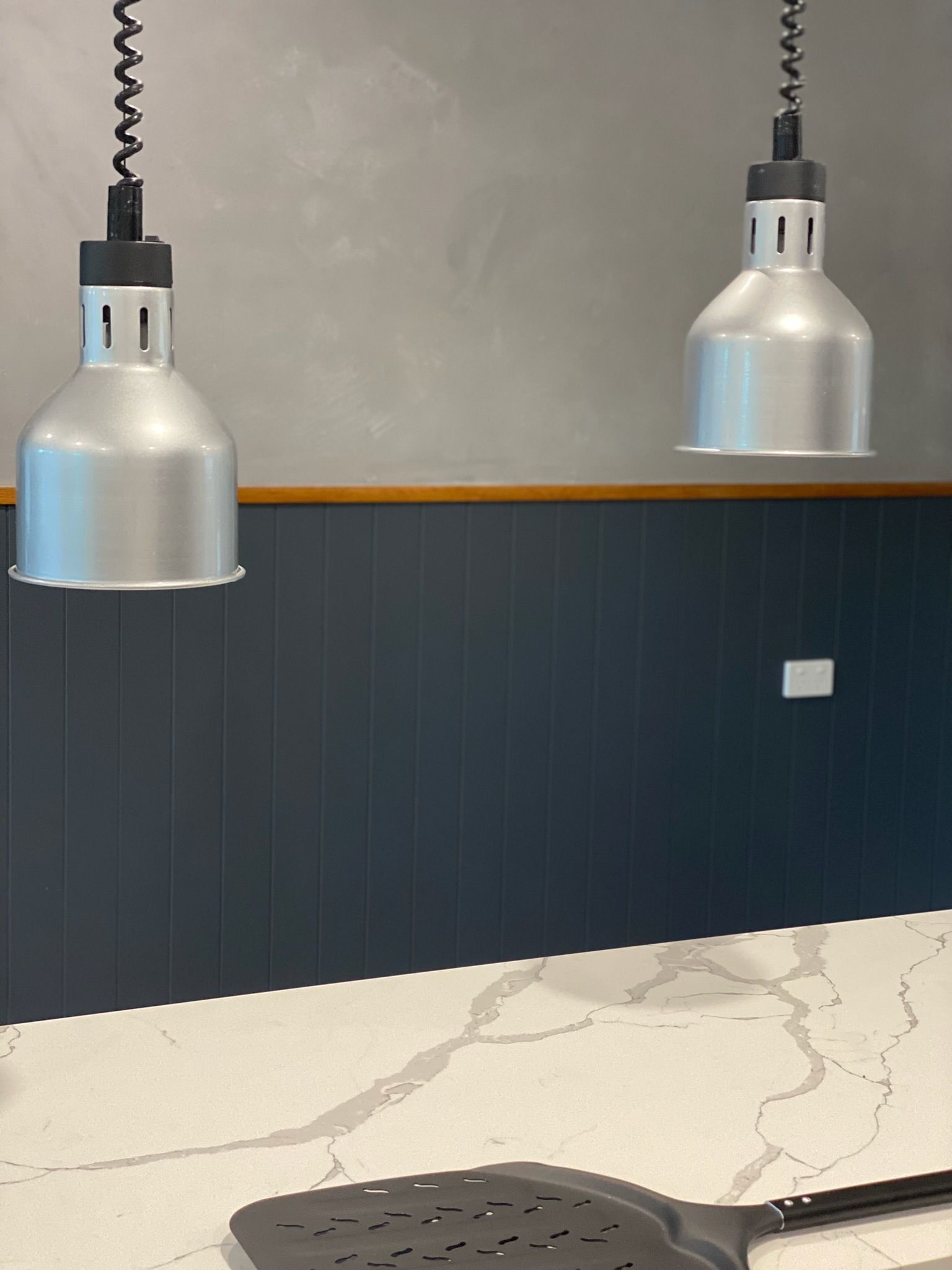 Two silver pendant lights over a marble counter with dark blue paneled wall behind them.