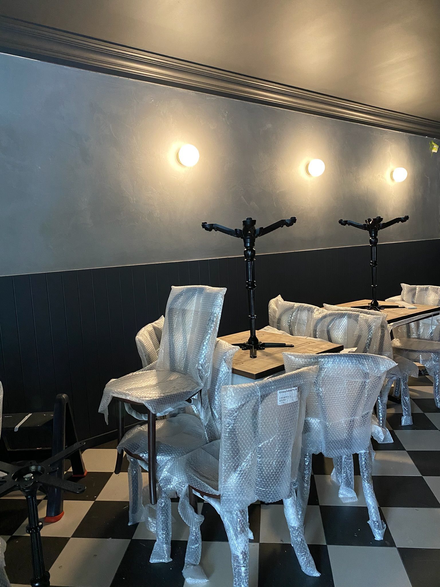 Small dining area with plastic-covered chairs and tables against a dark wall with wall lights
