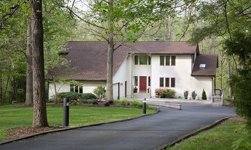 House with a dark driveway, trees, and a green lawn. The house is white with a brown roof.
