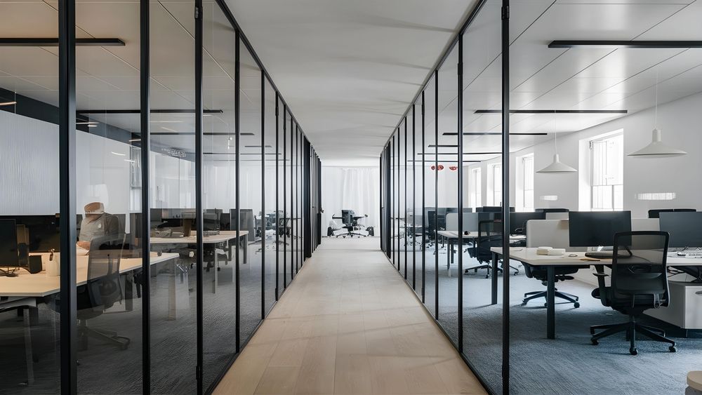Office interior with glass-walled workspaces lining a central hallway. Desks with computers and chairs are visible.