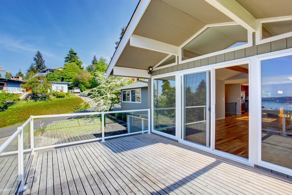 Wooden deck of a house with glass railing and sliding doors; view of greenery and water on a sunny day.