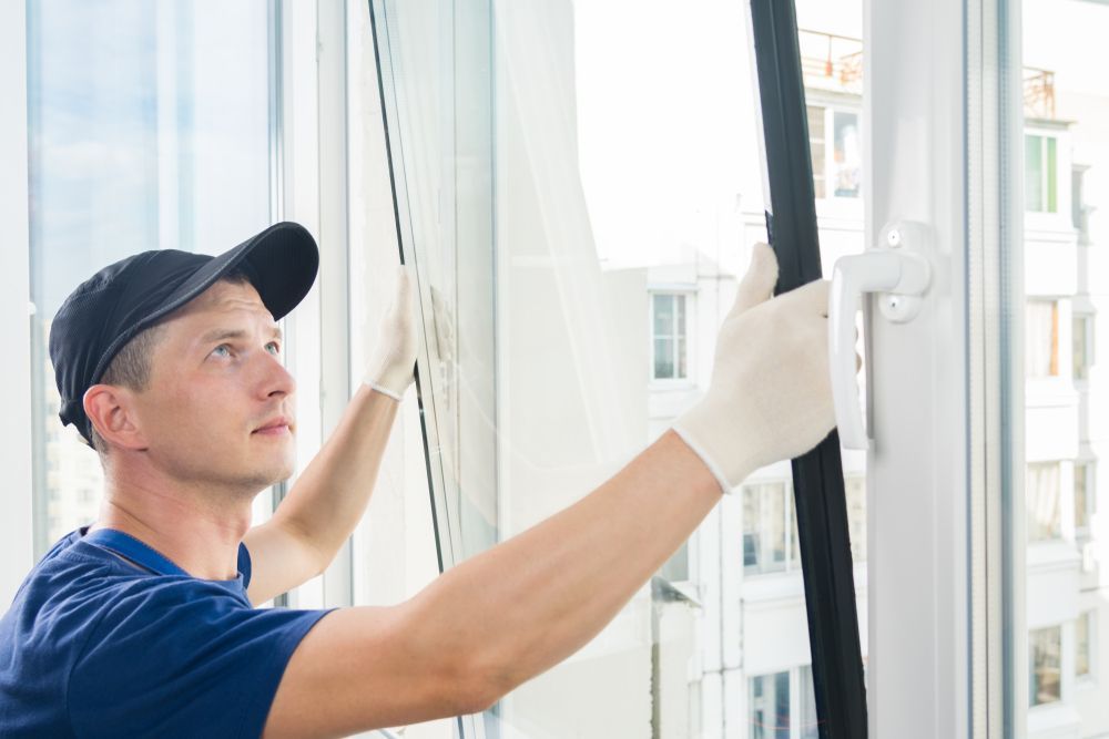 Man installing a window, wearing a cap and gloves, indoors with a bright background.