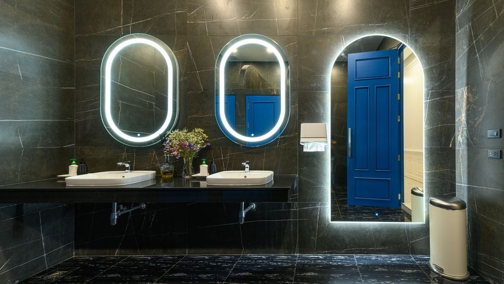 Public bathroom with dark marble walls, oval mirrors with lights, and blue stall doors.