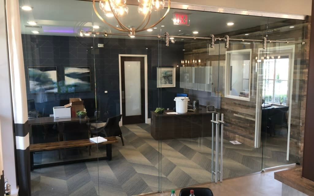 Office interior with glass walls and sliding door, desk, paintings, and chandelier.