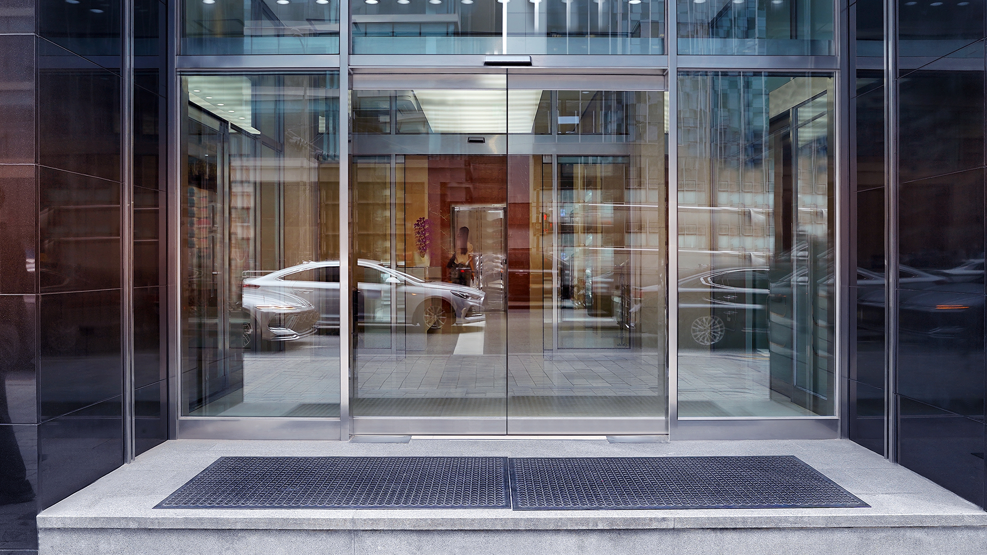Glass automatic doors of a building, reflecting a car and the interior. A black mat sits outside the doors.