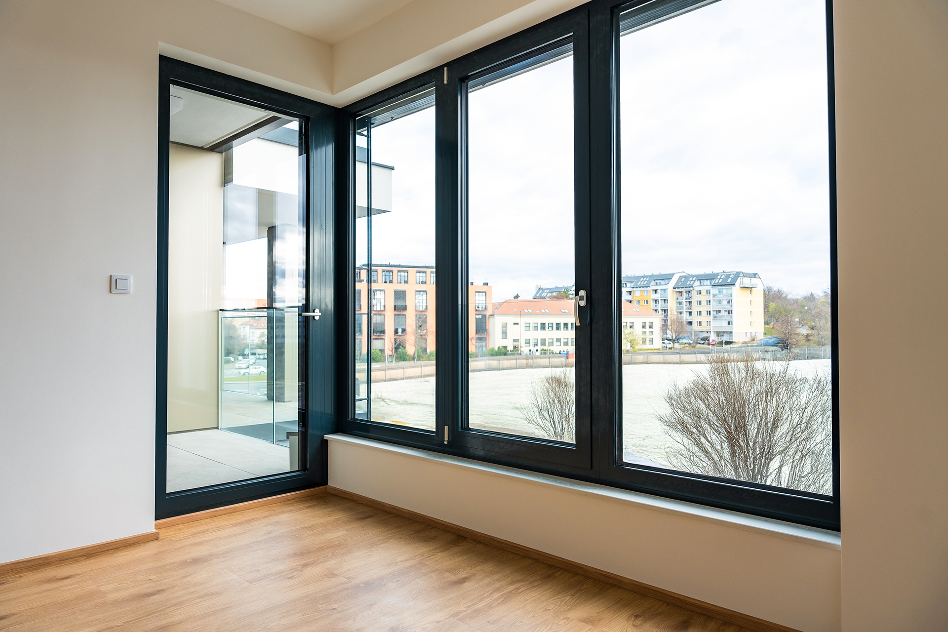 Bright room with large windows overlooking a city and a balcony. Wooden floor and white walls.