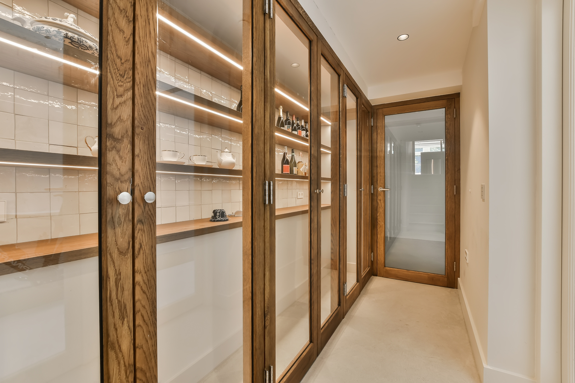 Wooden-framed glass pantry with shelves. Warm lighting and bottles on display in a narrow hallway.