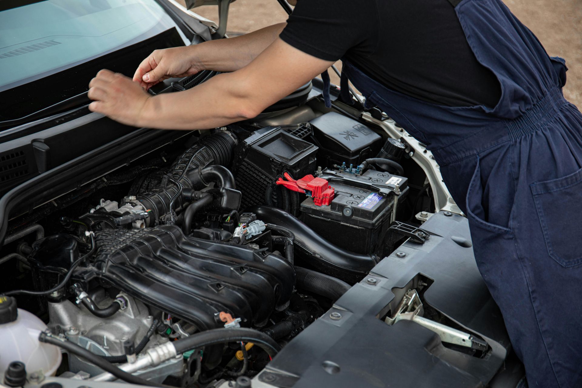 Person leaning over a car engine bay, inspecting the engine and battery.