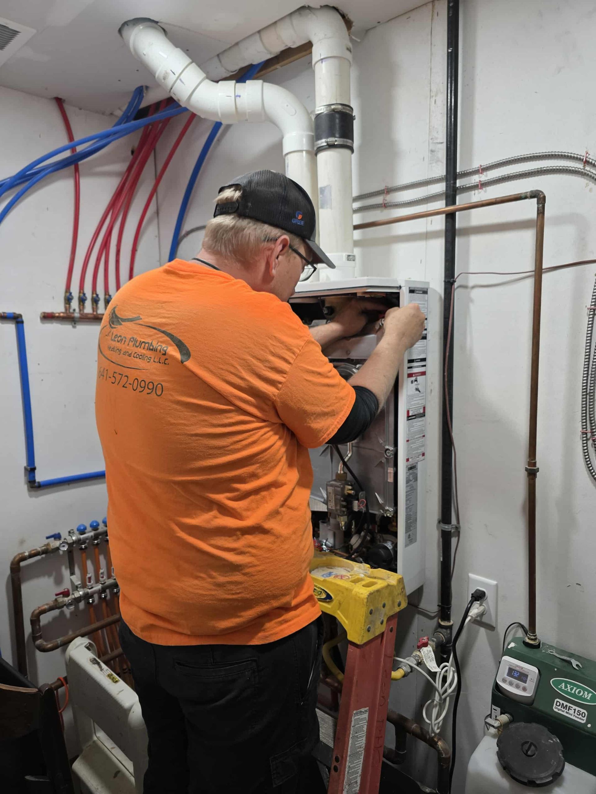 Man in orange shirt working on a wall-mounted appliance, plumbing visible in the room.