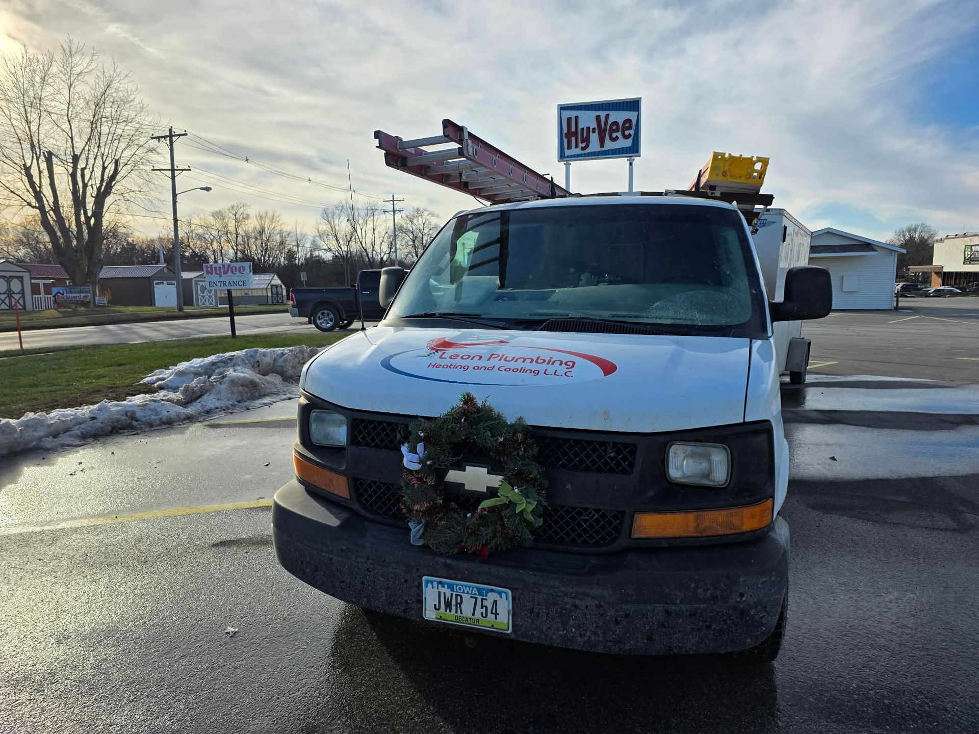 White work van parked in front of Hy-Vee grocery store with a ladder on top and a wreath on the front bumper.