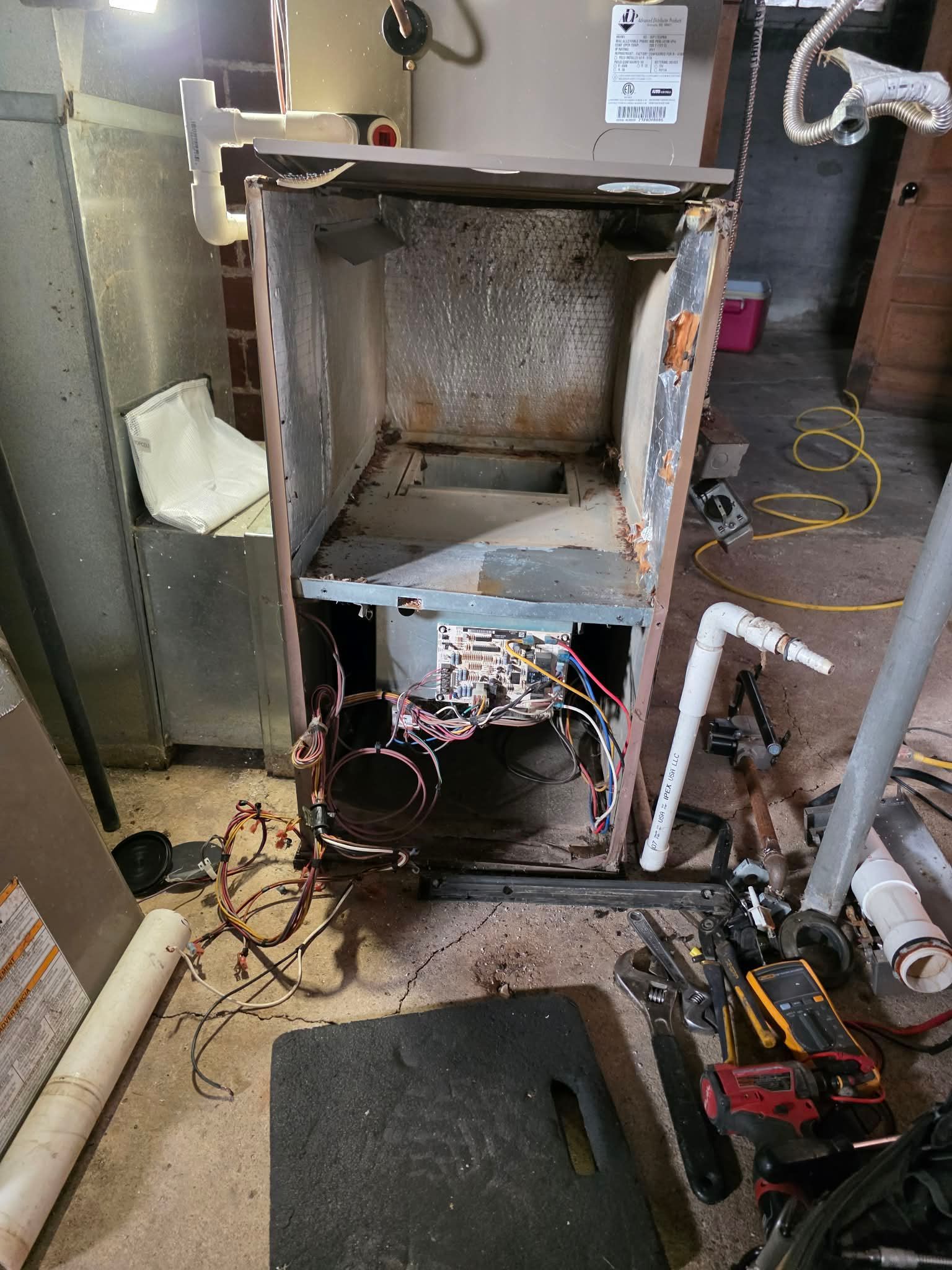 Open furnace with exposed components in a basement setting. Wires and tools are scattered around it.