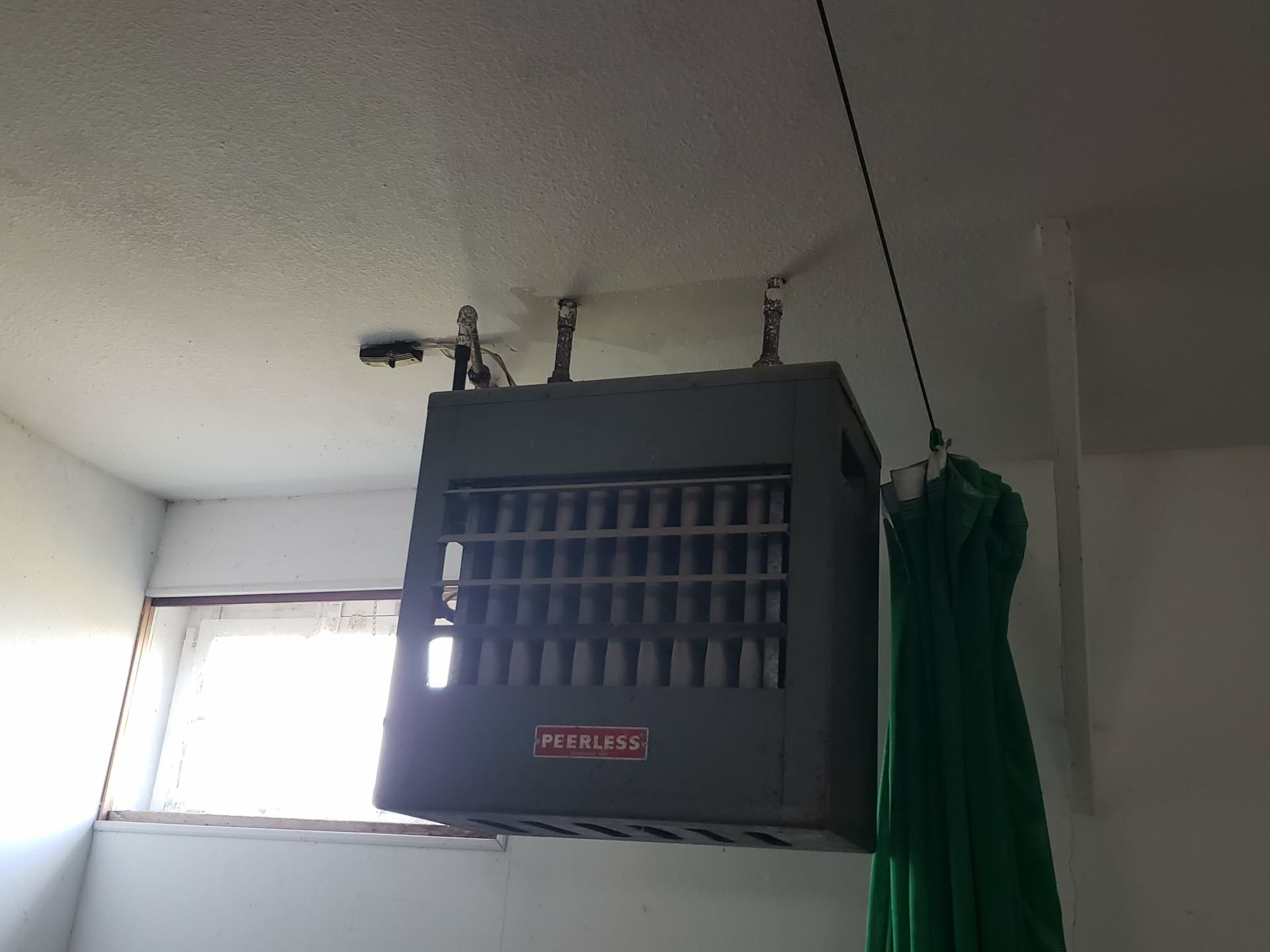 Ceiling-mounted Reznor gas heater in a room. Gray metal unit with a grill, hanging from the ceiling. Green curtain on right.