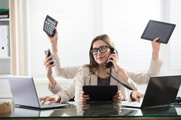 A woman is sitting at a desk with many hands holding a calculator , cell phone , tablet and laptop.