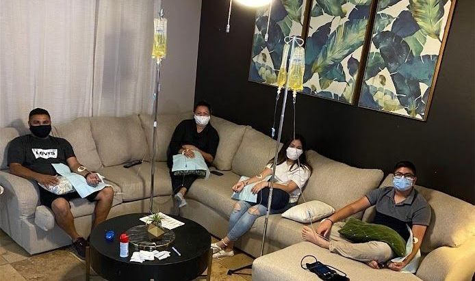 A group of people wearing face masks are sitting on a couch in a living room with IV.