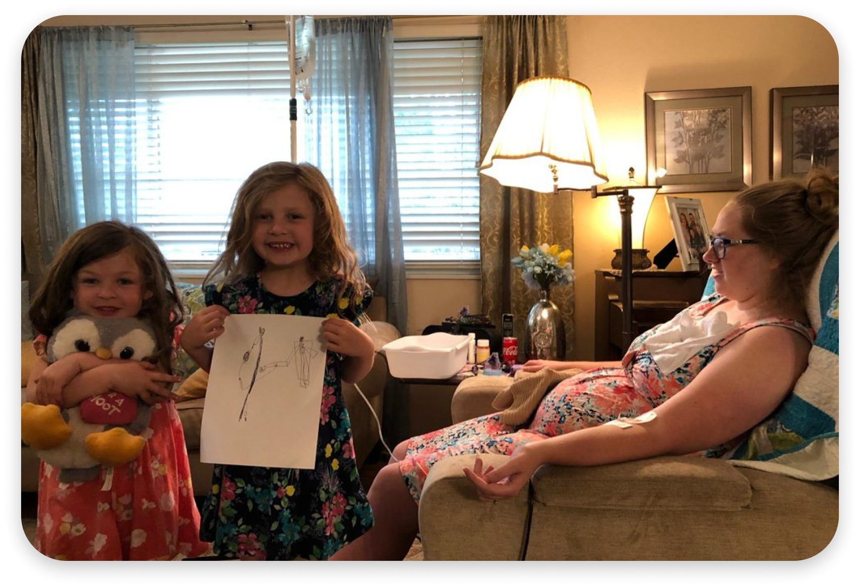 Two little girls are holding a drawing of a woman while a woman is sitting in a chair.