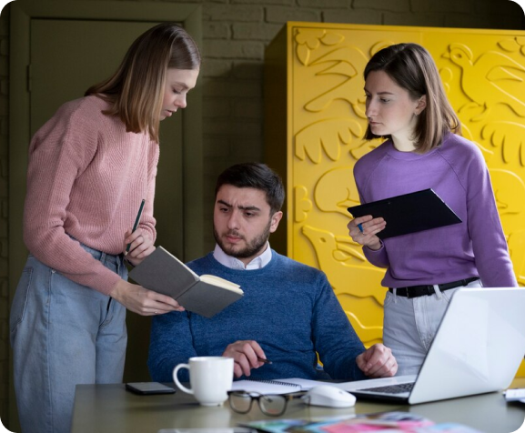 A group of people are standing around a table with a laptop.