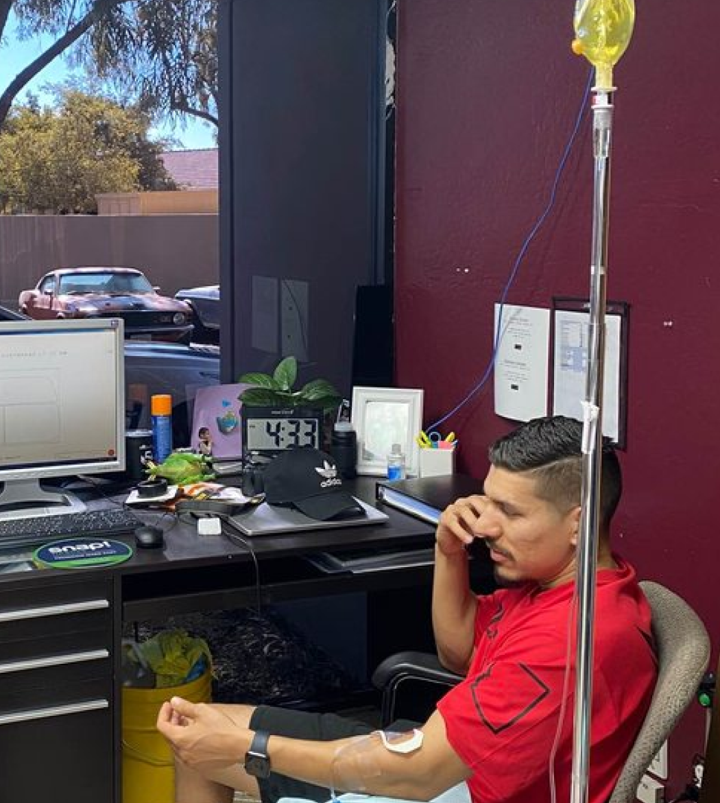 A man in a red shirt is sitting at a desk talking on a cell phone