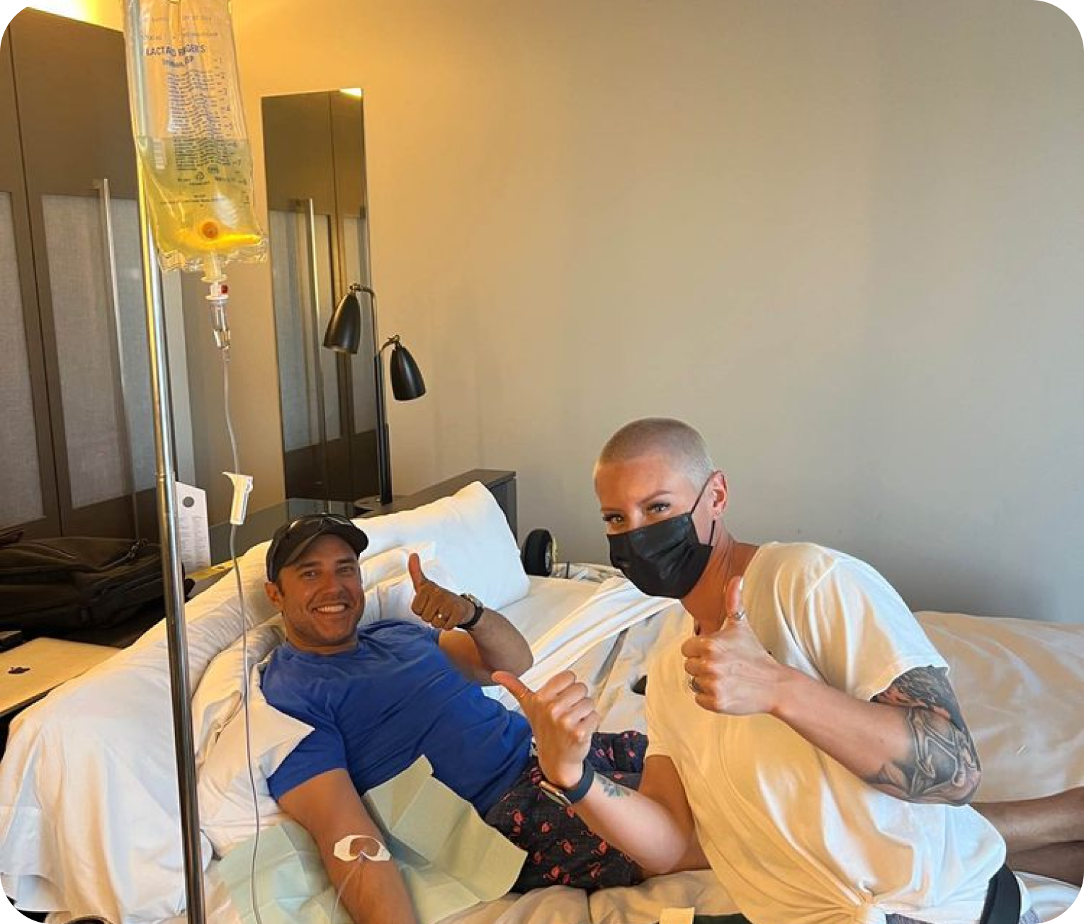 A man wearing a mask is giving a thumbs up next to another man in a hospital bed.