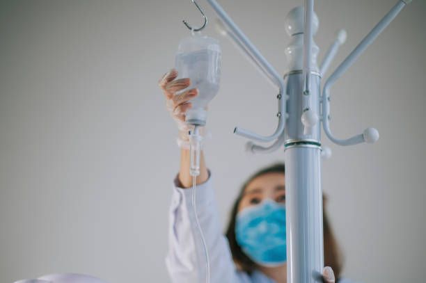 A nurse wearing a mask is holding an iv bag on a coat rack.