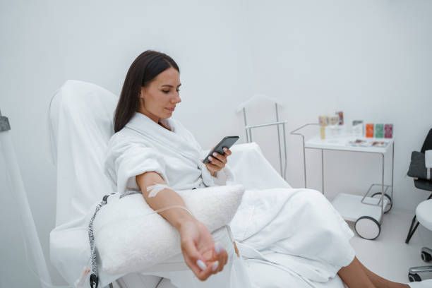 A woman is sitting in a chair with an iv in her arm and looking at her phone.