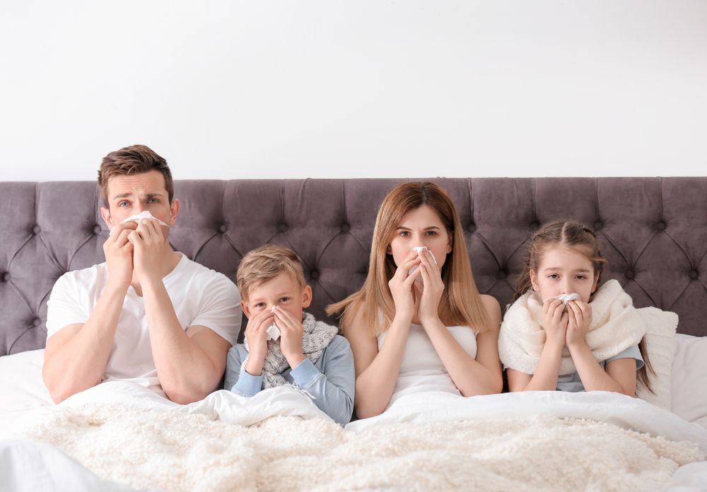 A family is sitting on a bed blowing their noses with tissues.