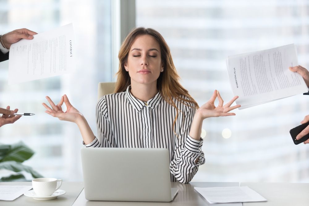 A woman is meditating while sitting at a desk in front of a laptop computer.