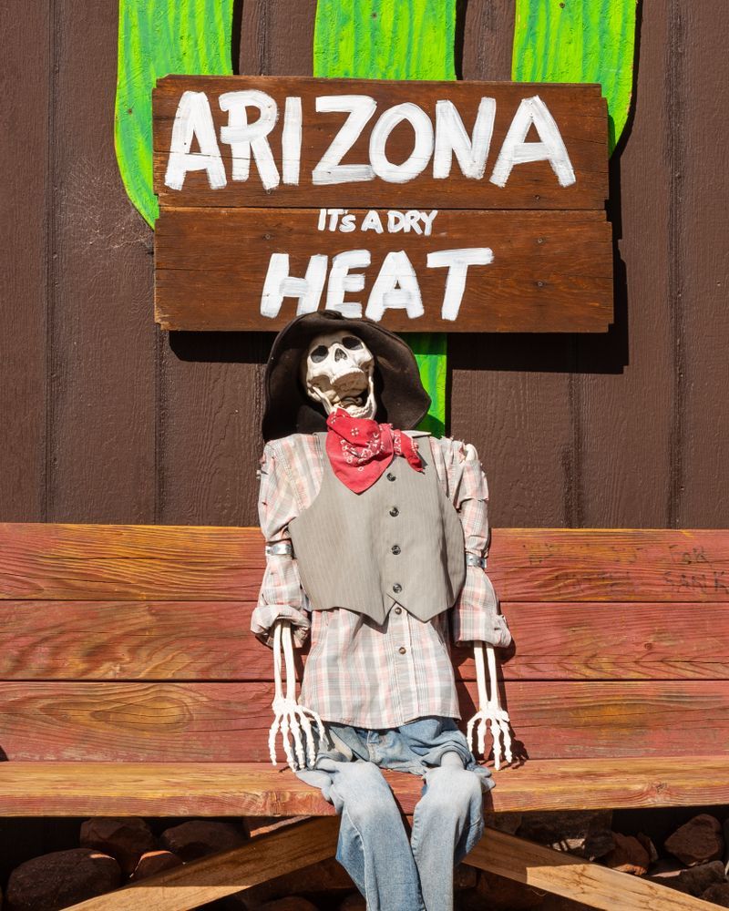 A skeleton is sitting on a bench under a sign that says arizona it 's a dry heat