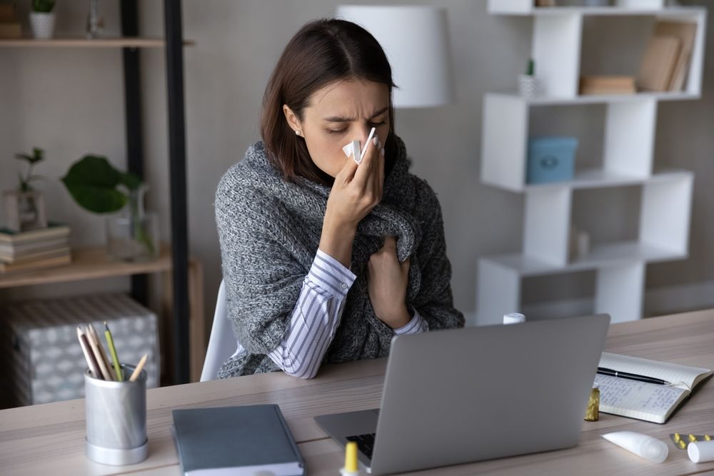 A woman is blowing her nose while sitting at a desk with a laptop.