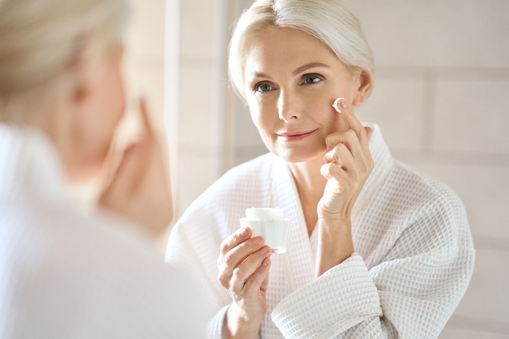 A woman is applying cream to her face in front of a mirror.