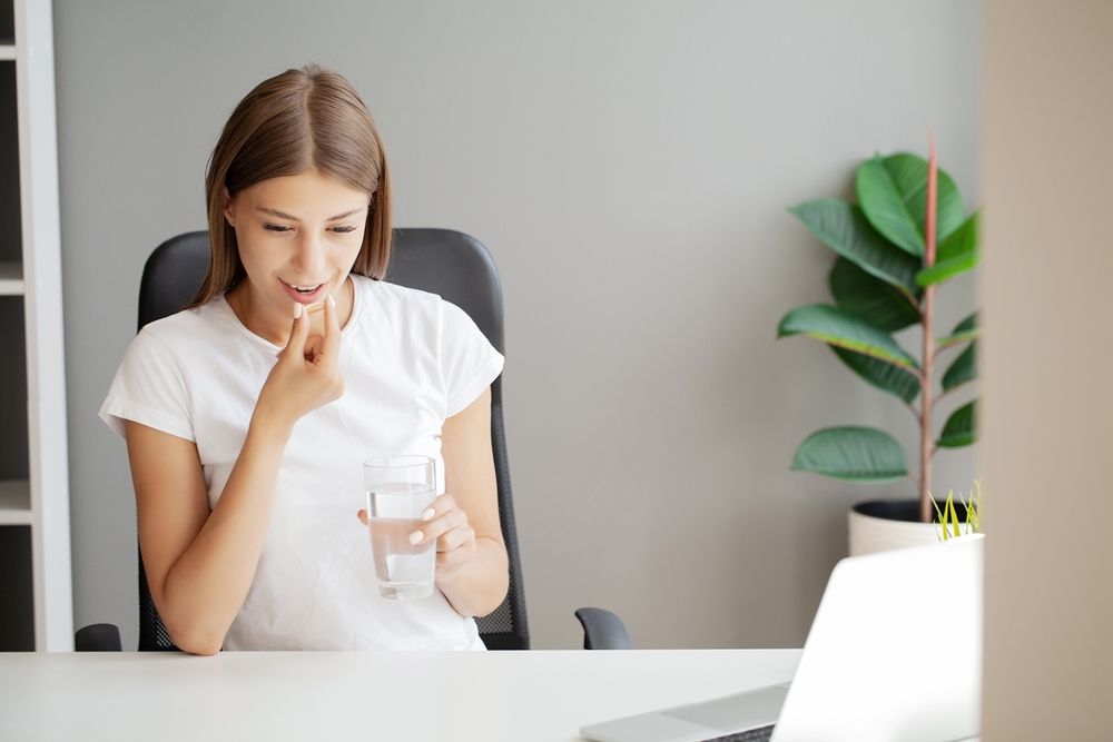 A woman is sitting at a desk taking a pill and drinking water.