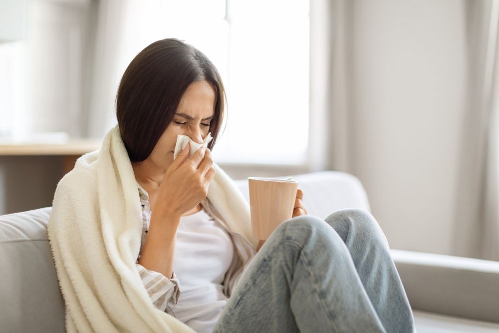 A woman is sitting on a couch blowing her nose while holding a cup of coffee.