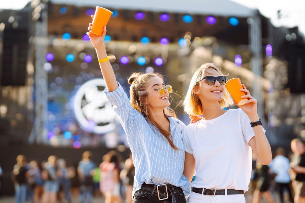 Two women are standing next to each other at a music festival holding cups in their hands.