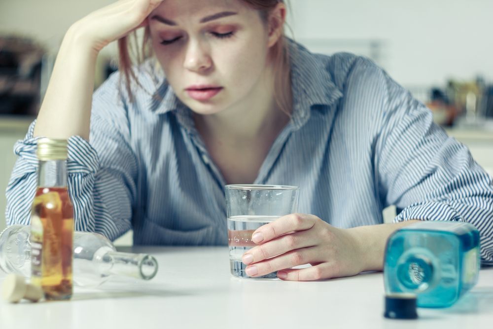 A woman is sitting at a table with a glass of water and bottles of alcohol.