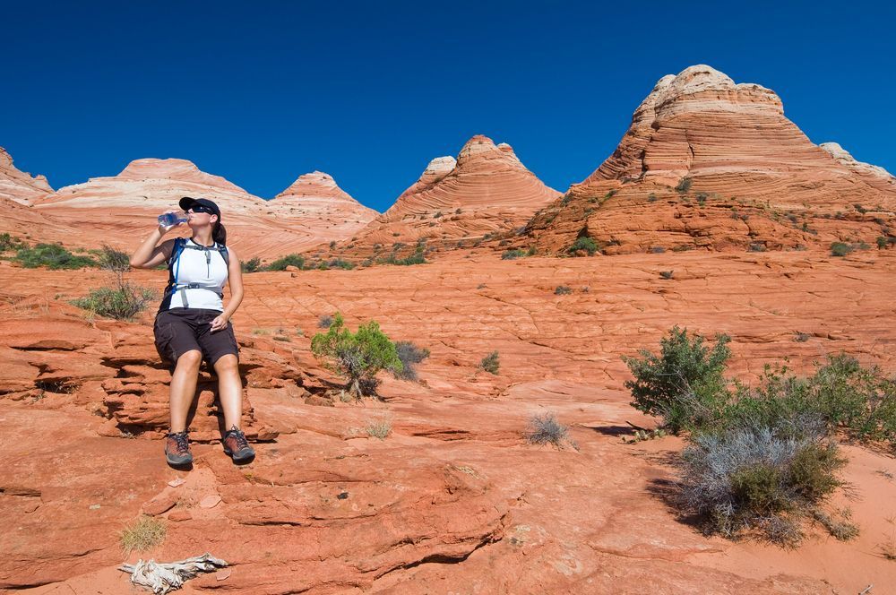 A woman is sitting on a rock drinking water in the desert.