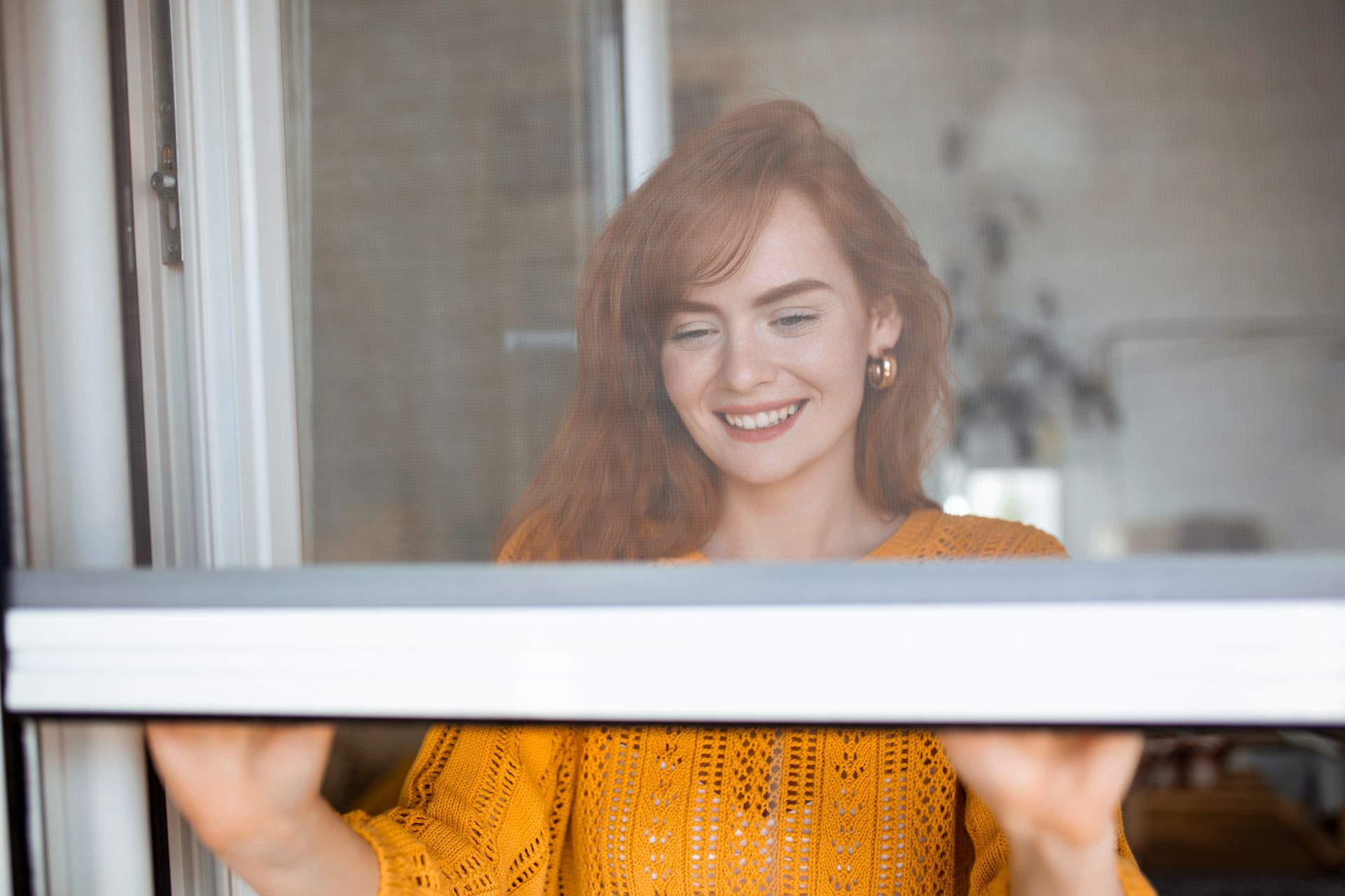 Una donna sorridente con un maglione giallo apre una finestra in casa.