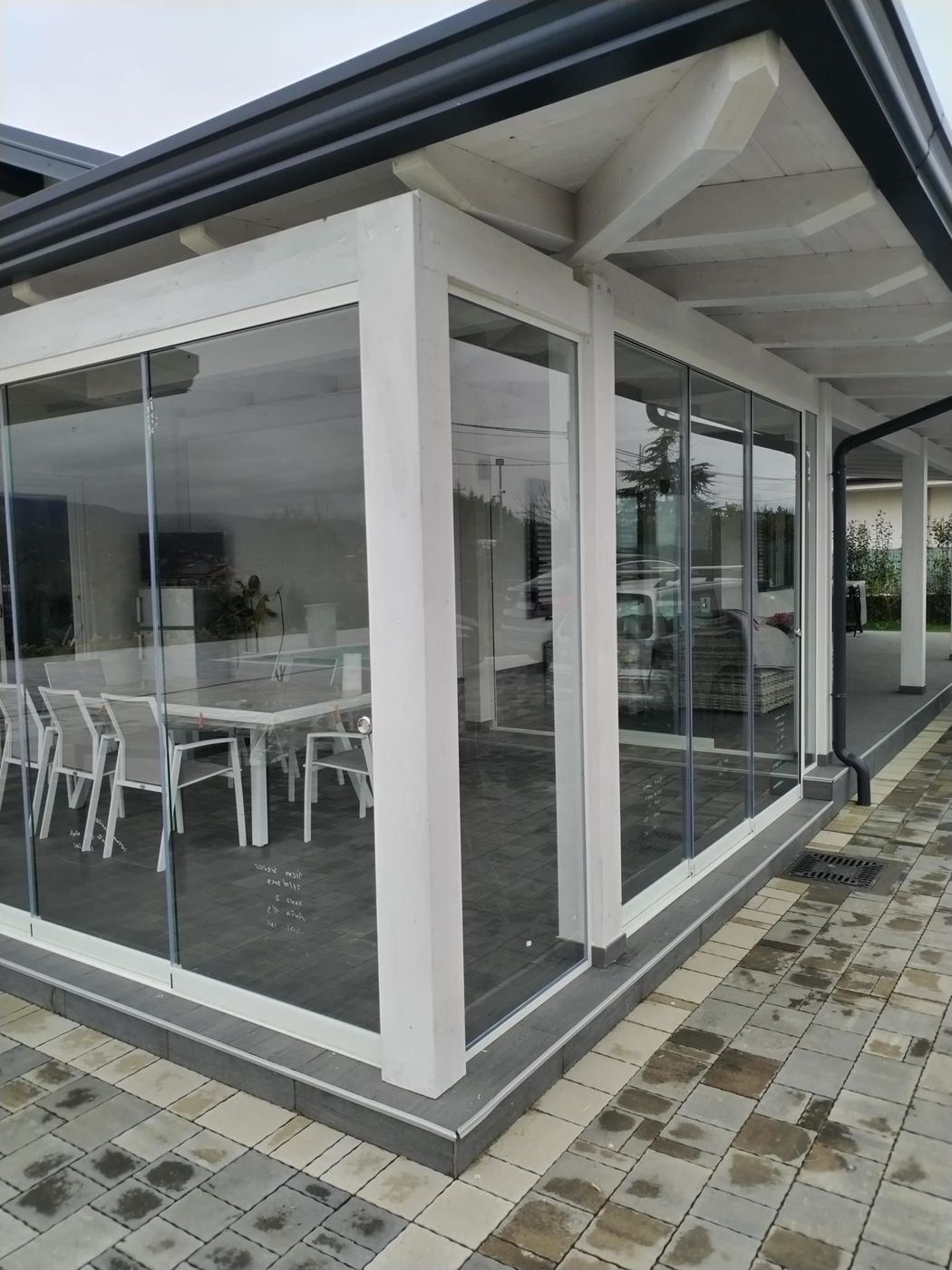 Area con tavolini e sedie bianche, separata da pareti in vetro, adiacente a un patio pavimentato e riparata da una tettoia.