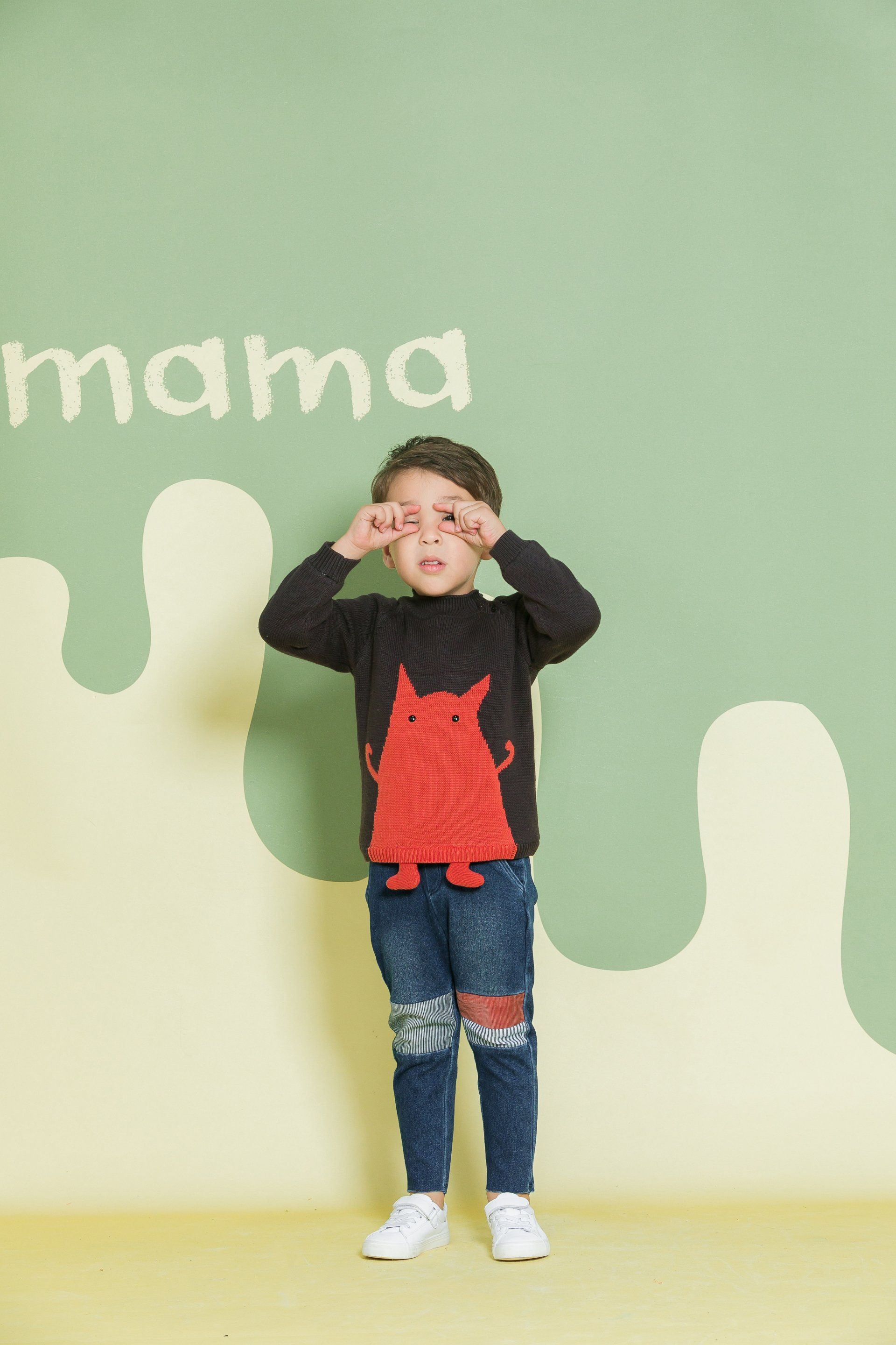 A young boy wearing a black sweater with a red monster on it is standing in front of a green wall.