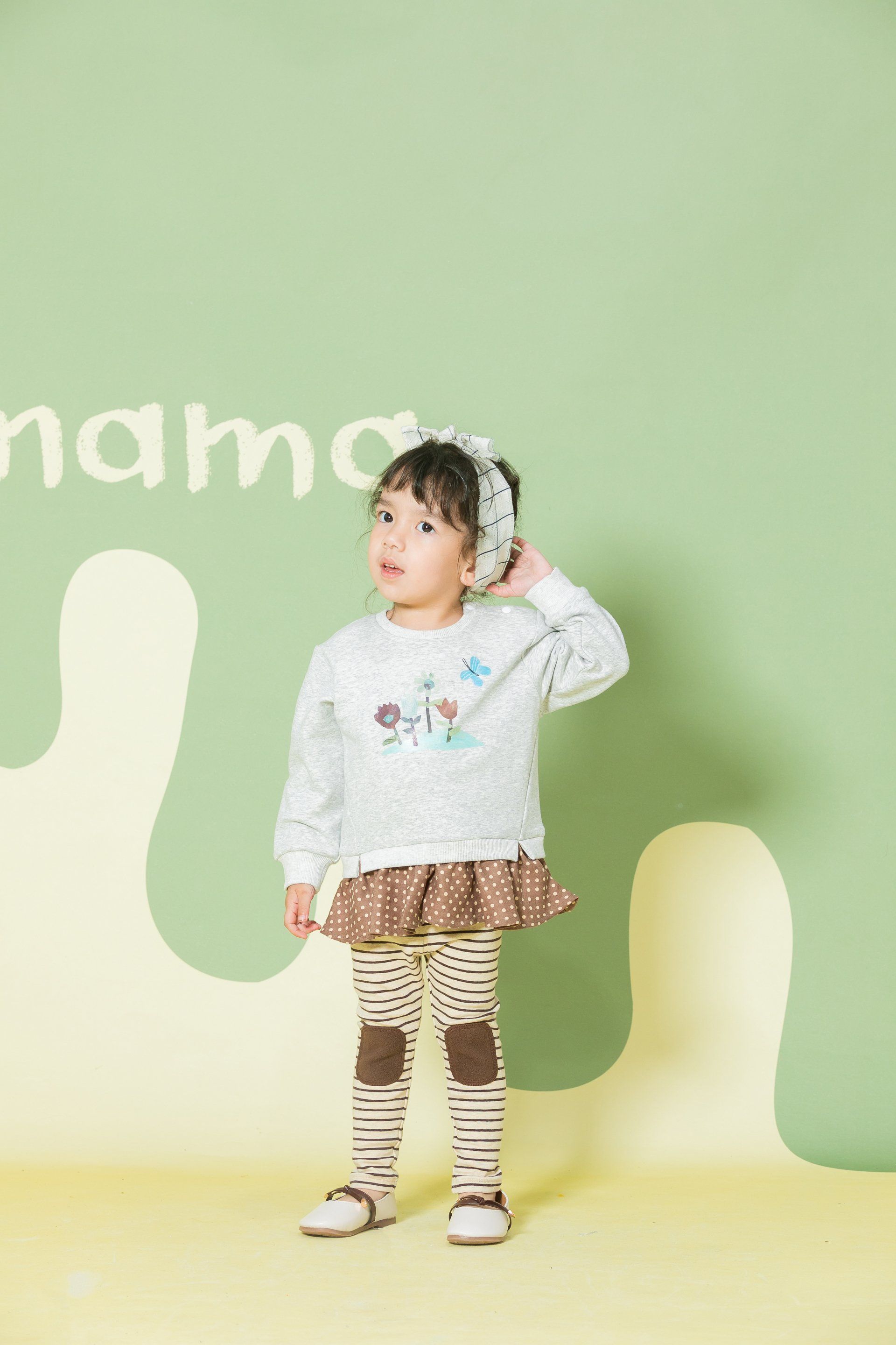 A little girl wearing a sweater and skirt is standing in front of a green wall.