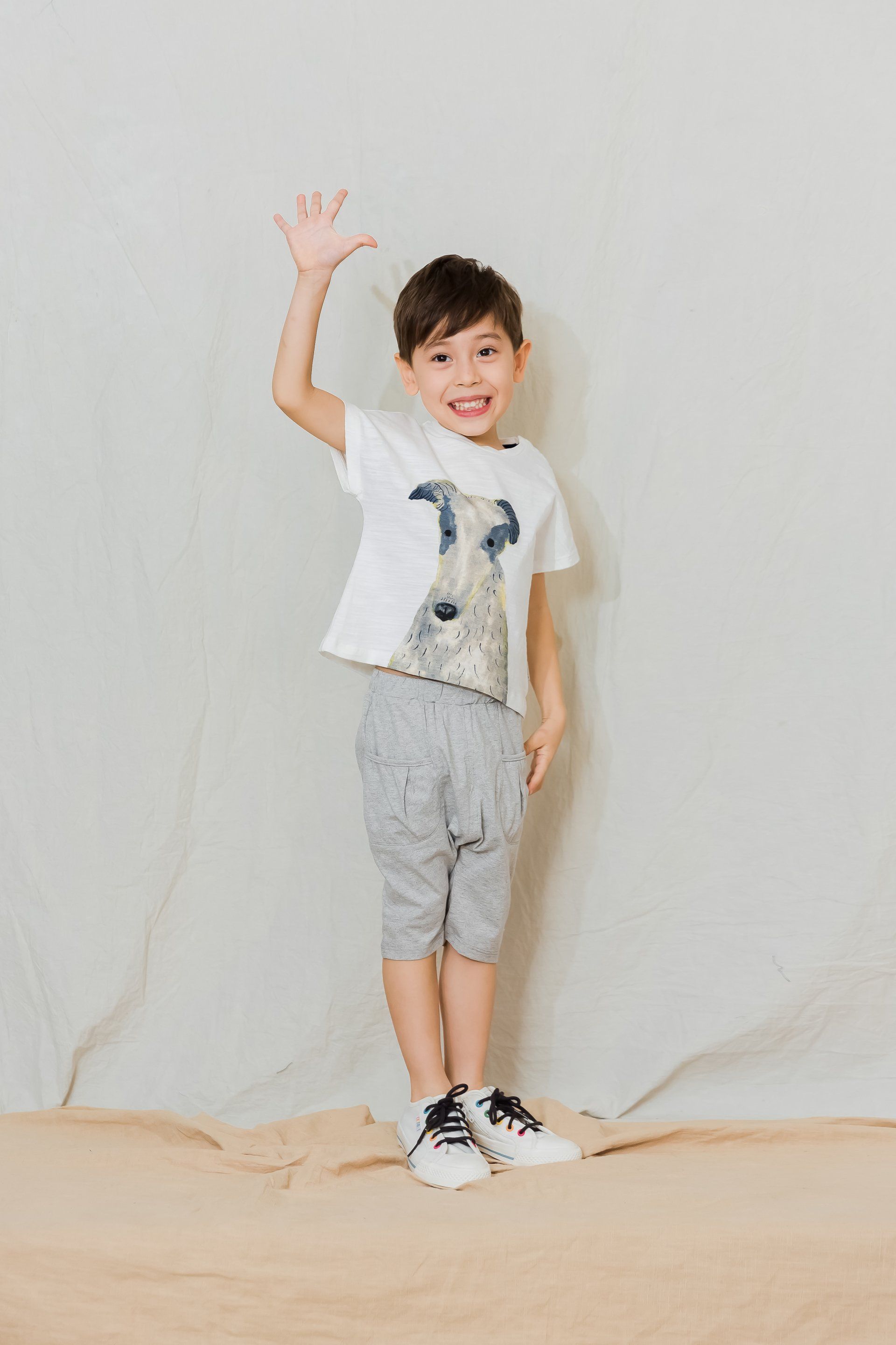 A young boy is standing in front of a white wall waving his hand.