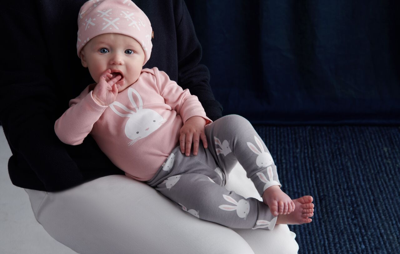 A baby wearing a pink hat and leggings is sitting on a pillow.