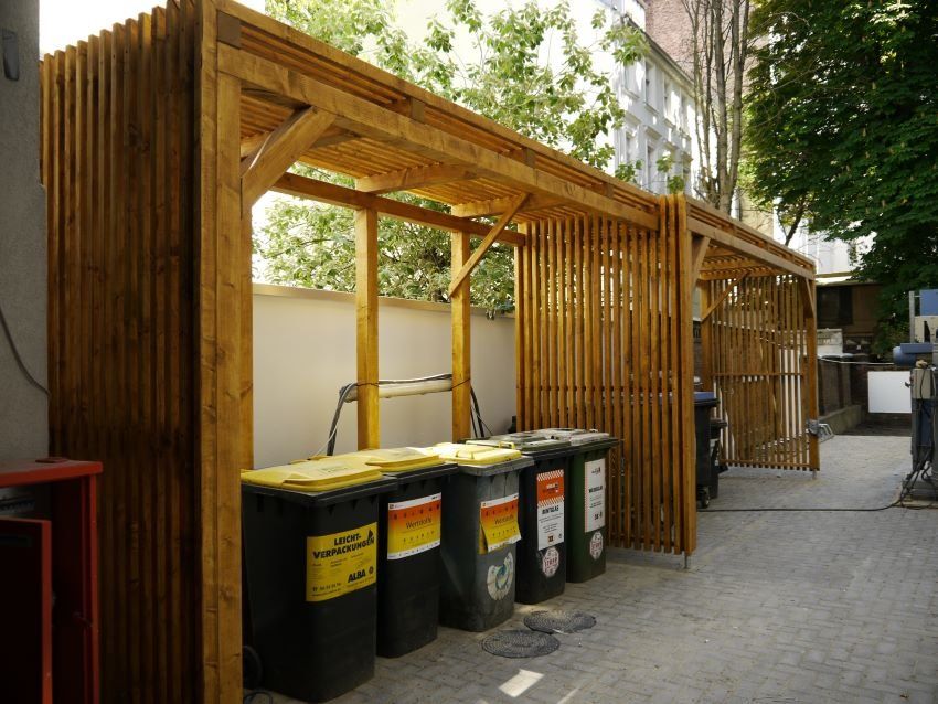 Unter einer Holzkonstruktion steht eine Reihe Mülltonnen.