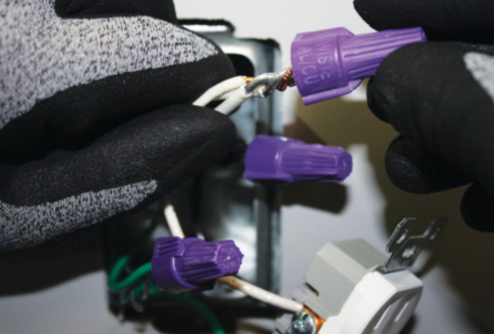 Gloved hands connecting wires with purple wire connectors in a white electrical box via pigtailing.