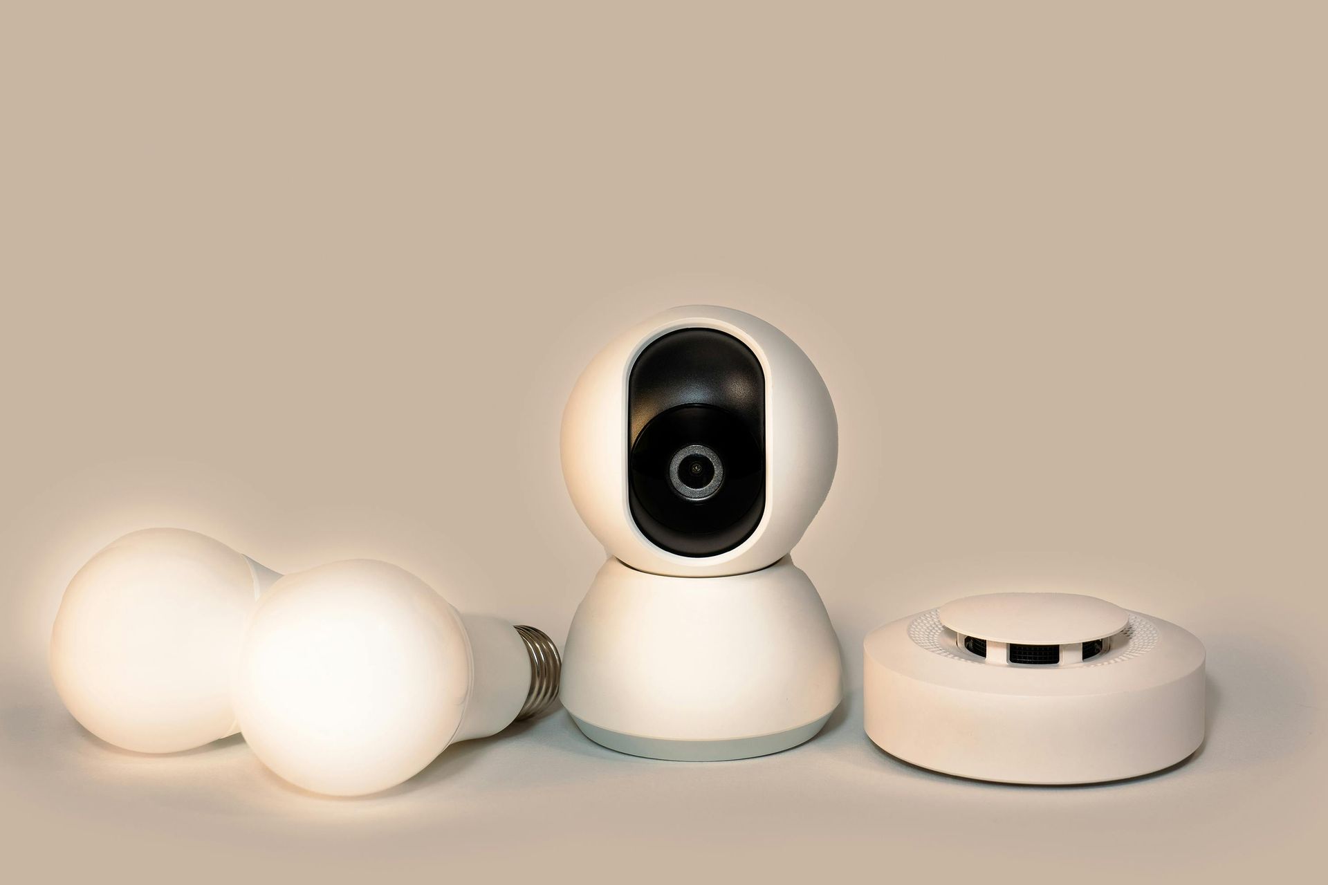 Smart home devices: light bulbs, security camera, and smoke detector on a beige surface.