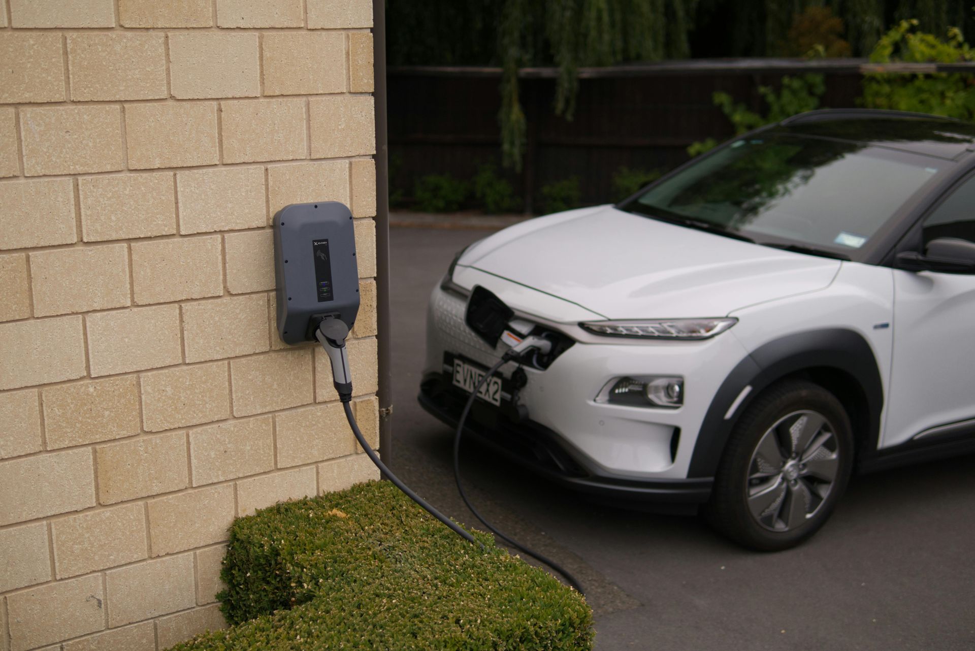 A white electric vehicle parked next to a brick wall, connected to a wall-mounted charging station.