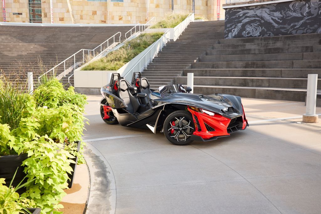 A red and black slingshot is driving down a street next to stairs.