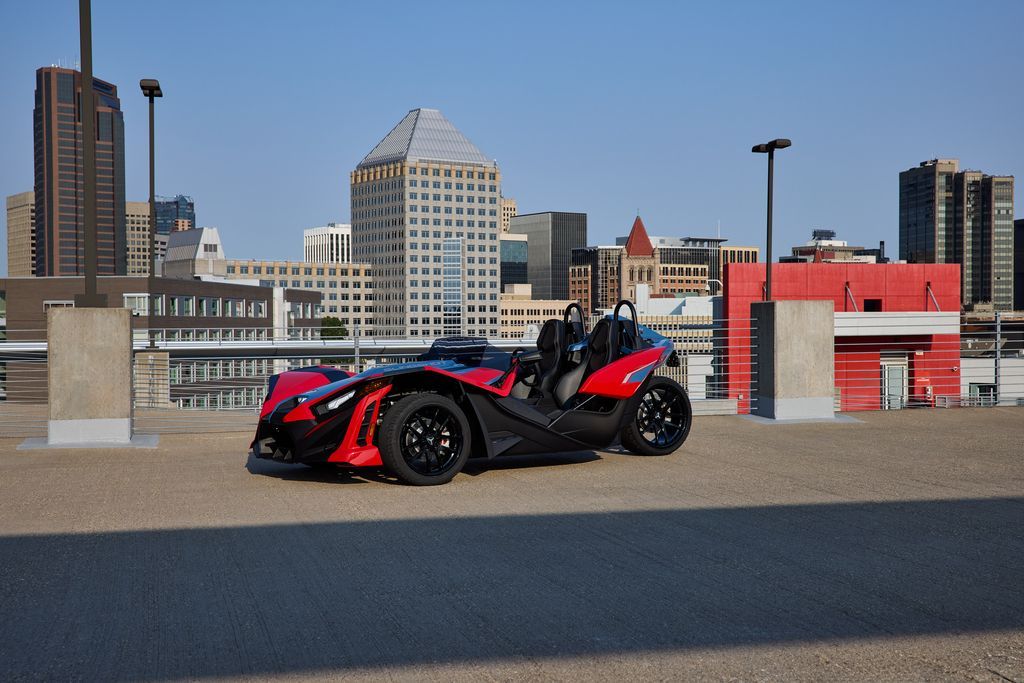 A red and black slingshot is parked on the side of the road in front of a city skyline.