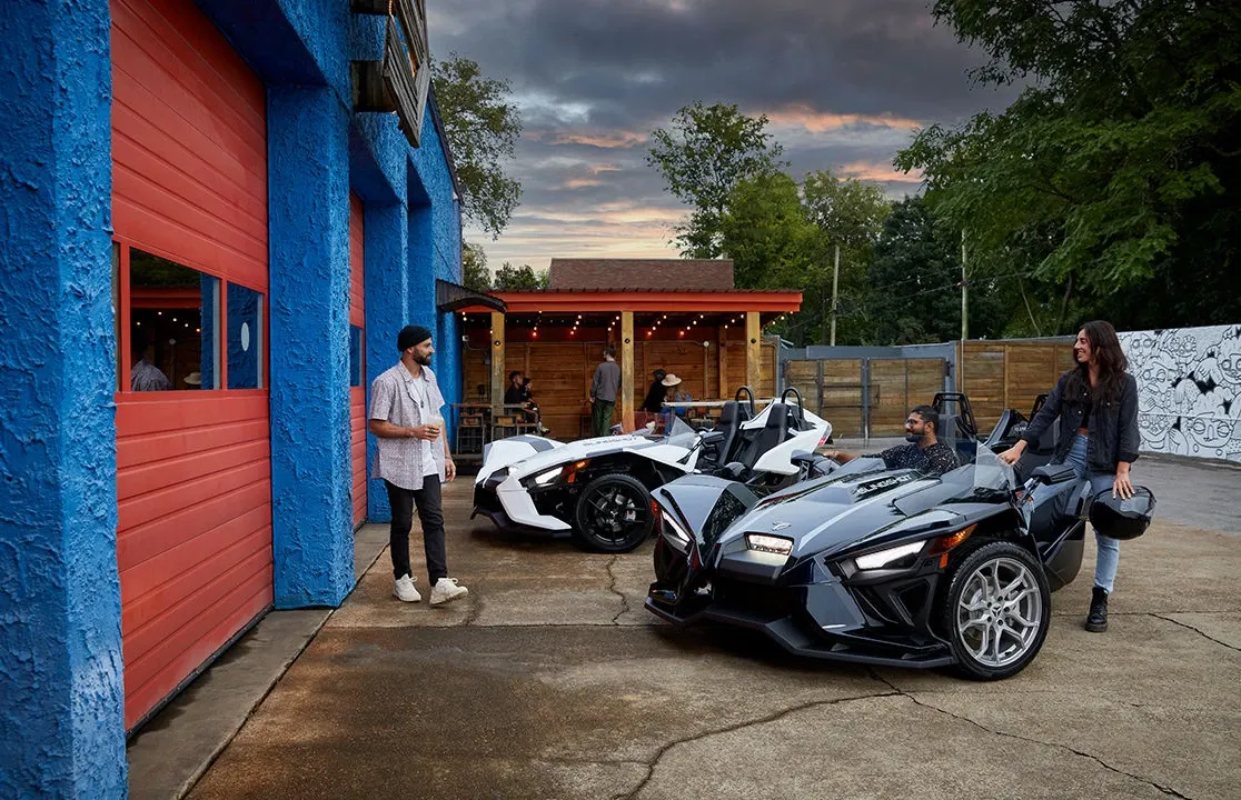 A man and a woman are standing next to a three wheeled vehicle in front of a garage.