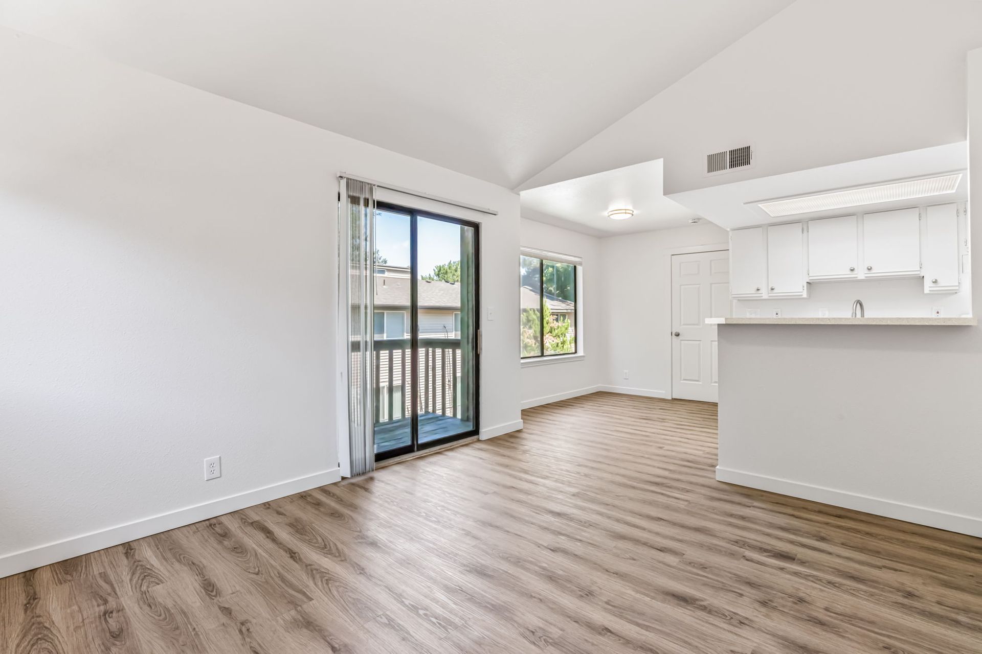Empty living room with wood-look flooring, balcony, and white kitchen counter.